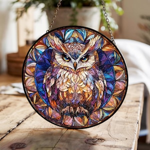 Puede incluir: Una ventana redonda de vidrieras con un diseño detallado de un búho. El búho tiene ojos amarillos y está rodeado por un colorido patrón de mosaico en tonos de azul, morado, naranja y amarillo. La ventana está enmarcada en negro y cuelga de una cadena.