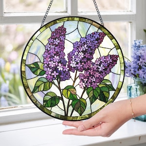 May include: A round stained glass window featuring a lilac flower design. The artwork showcases purple lilac blooms with green leaves and stems, set against a light blue and yellow background. The window is framed in black and hangs from a chain.