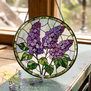 May include: A round stained glass window featuring a lilac flower design. The window has a black metal frame and a chain for hanging. The lilac flowers are purple with green leaves, set against a light green and white background.