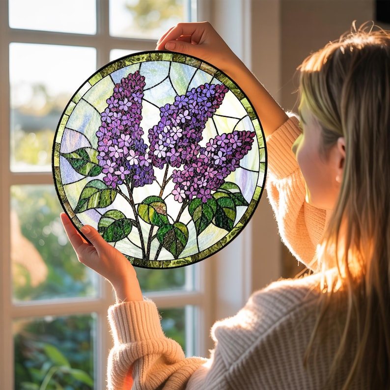 May include: A round stained glass window featuring a lilac flower design. The artwork showcases purple lilac blooms with green leaves and stems, set against a backdrop of various colored glass pieces. The window has a black border.
