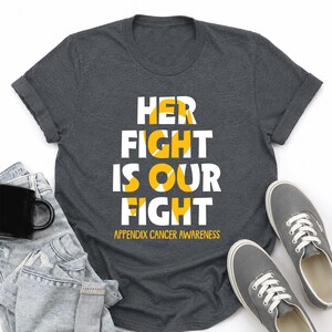 May include: A dark charcoal gray short-sleeve t-shirt featuring the white and yellow text "HER FIGHT IS OUR FIGHT" and "APPENDIX CANCER AWARENESS" with a yellow ribbon graphic integrated into the lettering on the front of the garment.