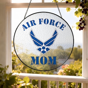 May include: Circular glass window hanging with the words "AIR FORCE MOM" in blue. The Air Force emblem is in the center. The glass has a textured finish and a black metal frame. The window hanging is suspended by a black chain.