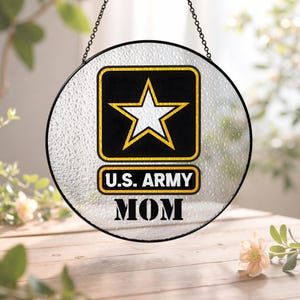 Op de afbeelding: Ronde glazen raamdecoratie met een zwarte rand en ketting om op te hangen. Het ontwerp toont het US Army-logo met een witte ster op een zwarte achtergrond en de tekst "U.S. ARMY MOM" in zwart en goud.