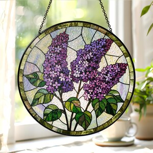 May include: A round stained glass window hanging featuring a lilac flower design. The artwork showcases purple lilac blooms with green leaves and stems, set against a light blue and white background. The window is framed with a black metal border and chain.