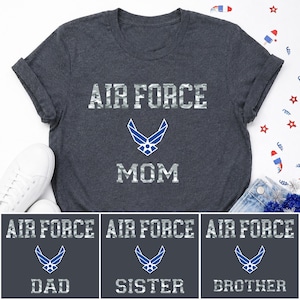 May include: A dark charcoal gray t-shirt featuring the text AIR FORCE in a white camouflage pattern, a blue and white Air Force emblem, and the word MOM in a white camouflage pattern. Additional text options include DAD, SISTER, or BROTHER.