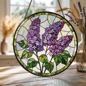 May include: Round stained glass window art featuring a lilac floral design. The artwork showcases purple lilac blooms with green leaves and stems, set against a light background. The piece is framed in black and hangs from a chain.