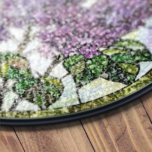 May include: Round floral patterned rug with a black border. The rug features a mosaic design with purple, green, white, and yellow colors. The rug is placed on a wooden surface.