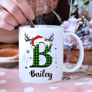 May include: White ceramic mug with a large green and black plaid letter "B" decorated with a Santa hat and antlers. The name "Bailey" is written in black script below the letter. Snowflakes and Christmas trees are also featured.