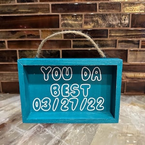 May include: A turquoise wooden sign with the words "YOU DA BEST" and the date "03/27/22" in white lettering. The sign has a jute rope handle and a rectangular shape.