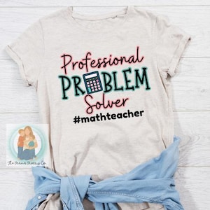 Puede incluir: Camiseta de color beige claro con el texto "Professional PROBLEM Solver" en rosa, verde azulado y negro, con un gráfico de calculadora. El hashtag #mathteacher está debajo. Una camisa vaquera azul está atada a la cintura.