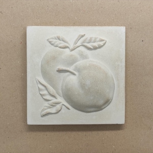 Puede incluir: Azulejo de relieve cuadrado blanquecino con dos manzanas y hojas. Las manzanas están texturizadas y tridimensionales, con sombreado sutil. Las hojas añaden un elemento naturalista al diseño.