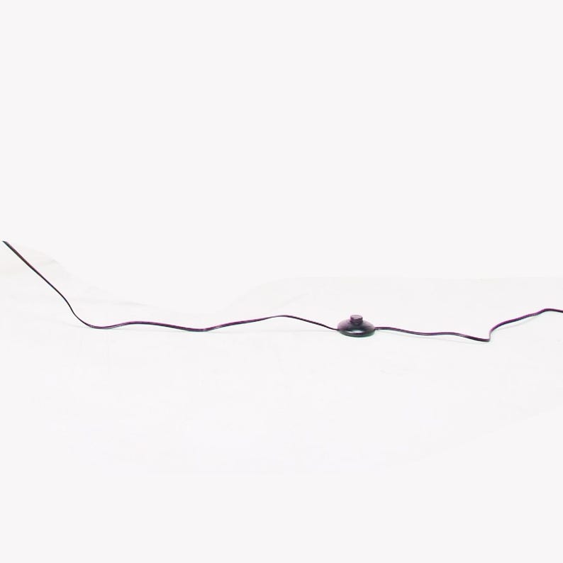 May include: A black electrical cord featuring a circular, flat, black floor switch positioned in the center. The cord extends horizontally across the frame, with the switch serving as a central control point for the connected electrical device.