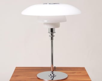 May include: A table lamp featuring a tiered design with three white glass shades of varying sizes. The lamp has a polished chrome metal stem and a circular chrome base. The structure supports the shades in a stacked arrangement, providing a clean, reflective surface on the metal components and a smooth, opaque finish on the glass elements.