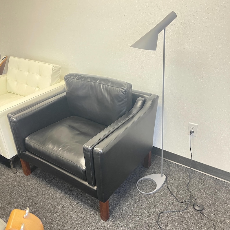 May include: A black leather armchair with a square back and seat cushion, supported by dark wooden legs. Beside it stands a tall, slender floor lamp with a matte gray conical shade, a thin metal stem, and a circular base with a power cord.