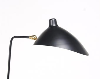 May include: A black metal lamp with a curved, dome-shaped shade and a slender black arm. The shade is connected to the arm by a polished gold-colored metal joint, allowing for an adjustable angle. The lamp features a smooth, matte finish.