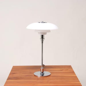 May include: A white, multi-tiered table lamp with a chrome base and stem. The lamp has a rounded shade and sits on a wooden table. The lamp's height is approximately 20 inches.