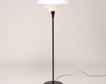 May include: A floor lamp featuring a thin black metal stand and a circular black base. The top consists of three stacked, tiered white glass shades of varying sizes that diffuse light, connected to a black cord extending from the base.