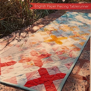 May include: A colorful English paper piecing table runner with a patchwork design. The table runner features a red cross pattern at one end, with yellow and pink cross patterns throughout. The text "Hot Cross Buns" is at the top.