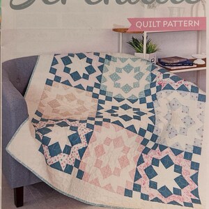 May include: A quilt pattern titled "Serenade" with a star design in shades of blue, pink, and white. The quilt is draped over a gray armchair. The quilt measures 56.5 inches by 56.5 inches.