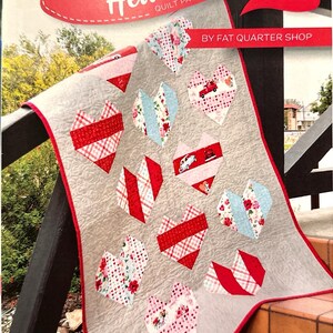 May include: A Heartstrings quilt pattern by Fat Quarter Shop featuring a gray quilt with red heart-shaped blocks in various patterns. The finished size is 32 1/2 inches by 44 1/2 inches. The design includes red, white, and blue floral and plaid fabrics.