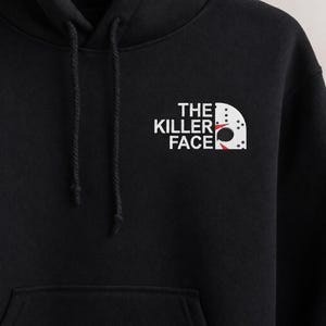 Könnte beinhalten: Schwarzer Kapuzenpullover mit weißem Text und einem grafischen Design. Der Text lautet "THE KILLER FACE" mit einer Hockeymaske. Der Hoodie hat einen Kordelzug und eine Vordertasche.