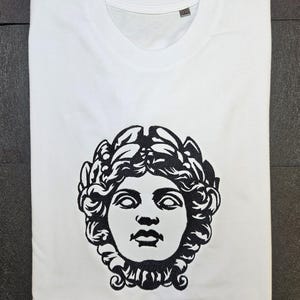 May include: White t-shirt with a black and white illustration of a classical bust wearing a laurel wreath. The t-shirt is neatly folded, highlighting the design. The bust design is centered on the front of the shirt.