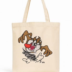 May include: Cream-colored tote bag featuring a cartoon illustration of the Tasmanian Devil. The character is depicted with brown fur, a red mouth, and a swirling tornado effect. The bag has two beige handles.