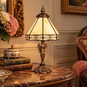 May include: A table lamp with a cream-colored, geometric stained glass shade and a bronze-toned base featuring a sculpted winged figure. The lamp stands on a dark, ornate pedestal base with intricate detailing, emitting a warm, soft light.
