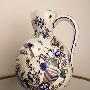 Könnte beinhalten: Eine weiße Keramikkrug mit bauchigem Körper und gebogenem Henkel. Der Krug ist mit farbenfrohen Blumen- und Vogelmotiven in Blau, Gelb, Rosa und Grün verziert. Der Boden und der Henkel sind blau eingefasst.
