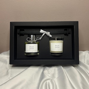 May include: A black gift set with a glass diffuser bottle and a candle, both labelled "Fresh Linen." The diffuser has a silver cap and reeds. A white ribbon ties the two items together. The candle is in a clear glass container.