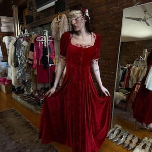 Può includere: Un lungo abito di velluto rosso con maniche corte e scollo quadrato. L'abito presenta un bordo in pizzo color crema intorno allo scollo e alle maniche. La modella indossa un copricapo abbinato con un accento floreale. L'abito è esposto in un negozio di abbigliamento vintage.