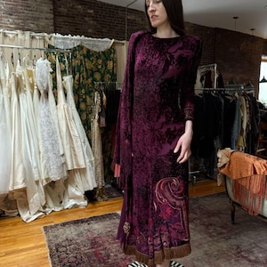 May include: A long, dark burgundy velvet dress with a floral pattern. The dress features long sleeves, a high neckline, and a decorative design near the hem. The dress has a fringe detail at the bottom.
