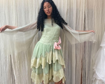 May include: A light green dress featuring long, sheer, flowing sleeves and a fitted bodice with ruched detailing. The skirt consists of multiple layered, ruffled tiers in varying shades of green, accented by a small, pale pink bow at the waist.
