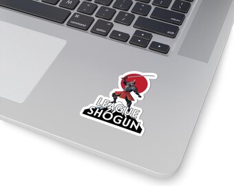 Può includere: Un adesivo con un guerriero samurai in piedi su una montagna con un sole rosso dietro di lui. Il testo "LEAGUE SHOGUN" è sotto il guerriero.