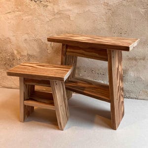 Könnte beinhalten: Zwei Holzhocker aus Naturholz mit rustikalem Finish. Der kleinere Hocker hat ein Regal unter dem Sitz. Der größere Hocker hat ein Regal unter dem Sitz und eine Rückenlehne.