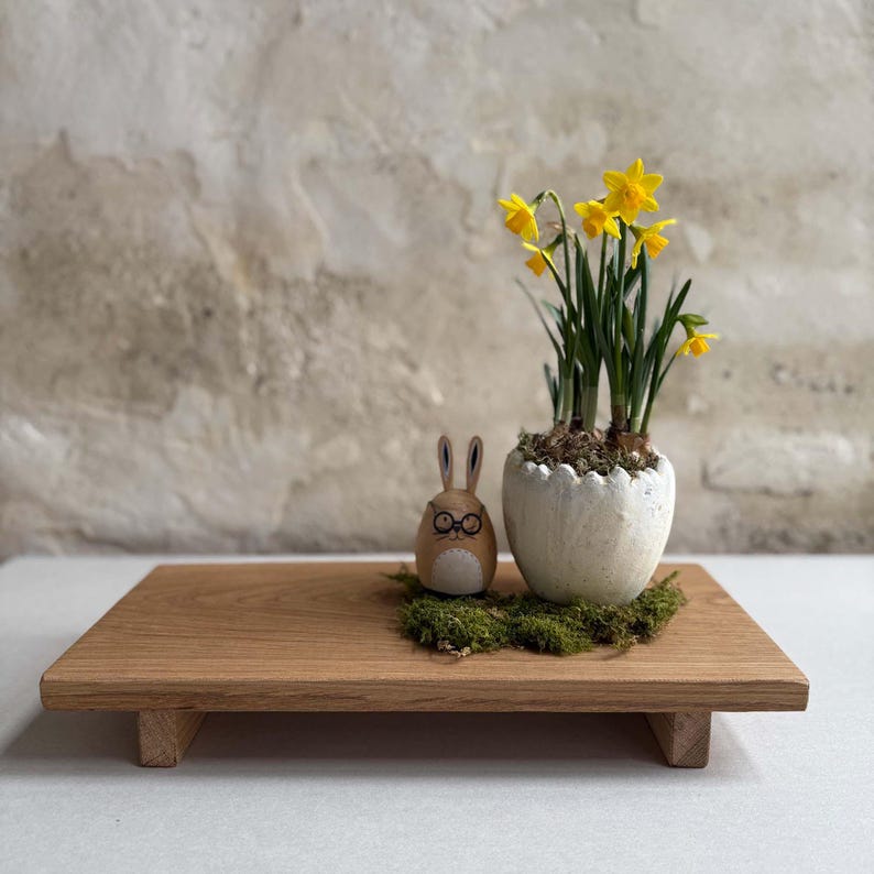 K&ouml;nnte beinhalten: Ein Holztablett pr&auml;sentiert eine Osterdekoration. Ein kleiner Holzhasen mit Brille sitzt neben einem wei&szlig;en, eif&ouml;rmigen Pflanzgef&auml;&szlig;, das mit gelben Narzissen gef&uuml;llt ist. Die Dekoration liegt auf gr&uuml;nem Moos.