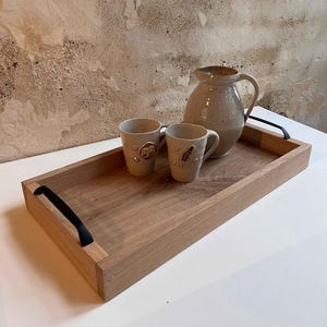 Könnte beinhalten: Ein Holztablett mit schwarzen Griffen, auf dem ein Keramik-Krug und zwei Tassen stehen. Der Krug und die Tassen sind mit einer weißen Glasur und braunen Designs verziert.