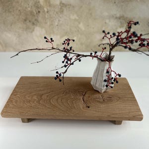 K&ouml;nnte beinhalten: Eine kleine wei&szlig;e Keramikvase mit strukturierter Oberfl&auml;che h&auml;lt einen Zweig mit roten Stielen und dunkelblauen Beeren. Die Vase steht auf einem rechteckigen Holztablett mit kleinen F&uuml;&szlig;en und zeigt eine nat&uuml;rliche Holzmaserung.