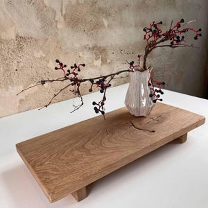 K&ouml;nnte beinhalten: Ein Holzserviertablett mit hellem Braunton, mit einer kleinen, wei&szlig;en, strukturierten Vase, die einen Zweig mit roten Stielen und dunklen Beeren h&auml;lt. Das Tablett ist rechteckig und ruht auf vier kurzen Beinen. Die Vase ist etwa 13 cm hoch.