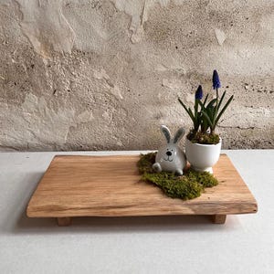 Könnte beinhalten: Ein Holztablett mit natürlicher Holzoptik. Das Tablett hat einen dekorativen Rand und zwei kleine Beine. Eine weiße Keramik-Hasenfigur und eine kleine Topfpflanze mit blauen Blüten befinden sich auf dem Tablett.