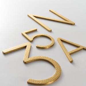 May include: Gold-coloured metal house numbers and letters with a hammered texture. The numbers are 5 and 7, and the letters are A and N. The numbers and letters are scattered on a white surface.