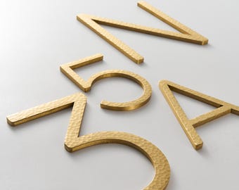 Könnte beinhalten: Goldfarbene Hausnummern und Buchstaben aus Metall. Die Zahlen und Buchstaben haben eine gehämmerten Textur. Die Zahlen sind 5 und 7, und die Buchstaben sind A und N. Die Zahlen und Buchstaben sind auf einer weißen Oberfläche verstreut.