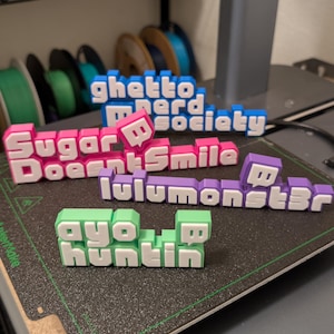 May include: 3D-printed text signs in various colors, including pink, blue, purple, and green, with white lettering. The signs feature phrases like "ghetto nerd society," "sugar doesn't smile," "lulumonstbr," and "ayo huntin." The items are on a black surface.