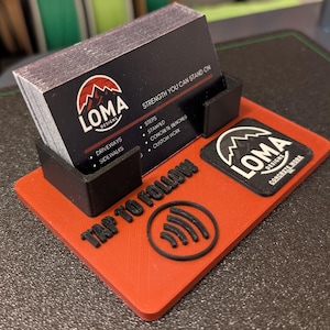 May include: A red business card holder with a black card holder and a stack of business cards. The cards have the "LOMA DESIGNS" logo and contact information. The base has the text "TAP TO FOLLOW" and a contactless payment symbol.