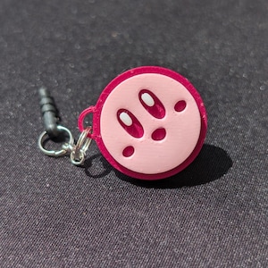 May include: A pink and red charm featuring a cartoon character face. The charm has a light pink face with dark pink eyes and mouth details. It includes a silver keyring and a black audio jack.