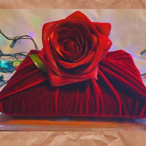 May include: A deep red velvet gift box is topped with a large, vibrant red rose. The rose has multiple layers of petals and a green leaf. The box is sitting on a wooden surface, with a brown paper background.