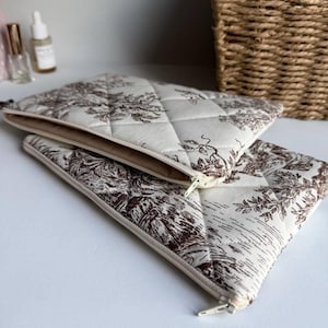 Puede incluir: Dos neceseres acolchados con un estampado toile de Jouy en marrón y crema. Las bolsas tienen forma rectangular con cierre de cremallera y diseño acolchado. Las bolsas están hechas de un material suave y acolchado.