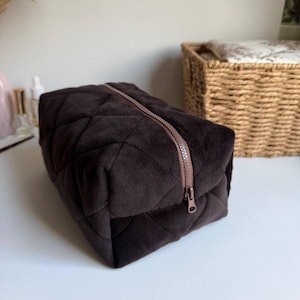 Puede incluir: Un neceser de terciopelo acolchado marrón oscuro con una cremallera marrón. La bolsa es rectangular y tiene un patrón de diamantes. La cremallera recorre la parte superior de la bolsa. La bolsa está sobre una superficie blanca.