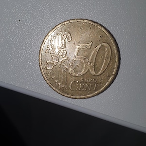Puede incluir: Una moneda de 50 céntimos de euro de color dorado con el mapa de la Unión Europea y estrellas en un lado. El otro lado muestra el número 50 y las palabras "EURO CENT".