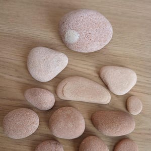 Pode incluir: Uma coleção de onze pedras lisas e arredondadas em vários tons de rosa seco e terracota claro. As pedras variam em tamanho e forma, sendo que uma pedra maior apresenta uma pequena mancha branca circular na sua superfície texturizada.